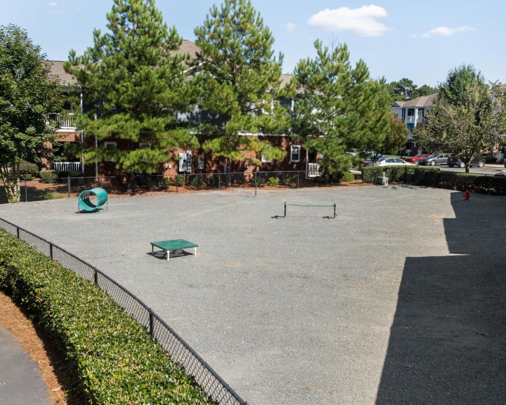 Fenced dog park with gravel ground, agility equipment, and trees, surrounded by apartment buildings.